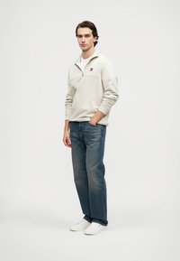 Maglione zip-up beige chiaro con un piccolo logo, abbinato a jeans blu medio e sneakers bianche. Il maglione ha maniche lunghe e una vestibilità comoda.