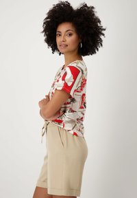 Chemise à manches courtes en tissu léger arborant un motif rouge, blanc et beige avec divers éléments graphiques. Associée à un short beige clair.