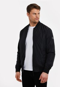 Schwarze Bomberjacke mit glatter Textur, Reißverschluss, gerippten Bündchen und einem kleinen Emblem auf dem linken Ärmel. Getragen über einem weißen Hemd.
