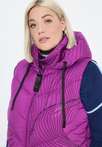 Fialová péřová vesta s kapucí, se černými liniovými vzory, předním zipem a stahovací šňůrkou. Nositá přes modrý, texturovaný svetr.