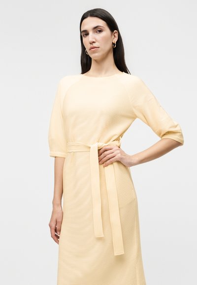 Γυναίκα που φοράει ένα ανοιχτό κίτρινο ριμπ φόρεμα με μανίκια μήκους ως τον αγκώνα και ζώνη στη μέση που ταιριάζει, ποζάροντας με το ένα χέρι στη μέση.