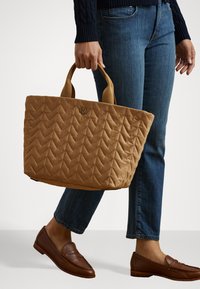 Quilted tan tote bag with chevron pattern, fabric handles, and R.L. logo. Paired with blue jeans and brown loafers.