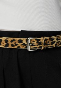 Pasek w panterkę wykonany z teksturowanej tkaniny z srebrną metalową klamrą. Noszony na czarnych spodniach, z kilkoma otworami do regulacji.