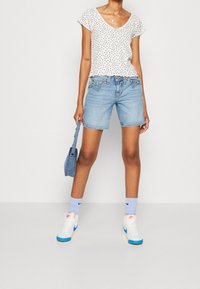 Top blanco de manga corta con estampado floral negro, pantalones cortos de mezclilla azul, zapatillas altas blancas con acentos azules, llevando una pequeña bolsa de mezclilla azul.