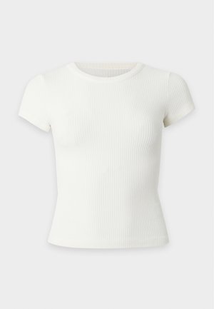 Ung kvinde med langt sort hår iført en tætsiddende hvid cropped top, blå bukser med vide ben og sort bælte samt sorte spidse sko, stående mod en hvid baggrund.