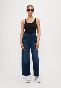 Svart tanktopp, høy midje, vide blå jeans og hvite joggesko. Modell iført svarte solbriller. Glatt tekstur, uformell stil.