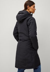 Lang zwarte winterjas met een capuchon, voorzien van een getailleerde taille en textuurstof. Inclusief zijzakken en minimalistische ontwerpelementen.