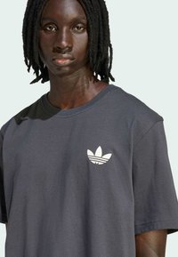 Tmavosivá bavlnená tričko s okrúhlym výstrihom a krátkymi rukávmi, so bielym logom Adidas na ľavej strane hrudníka. Jemná textúra.