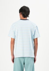 En mann står med ryggen til kameraet iført en lyseblå stripete t-skjorte og teal shorts mot en enkel hvit bakgrunn.