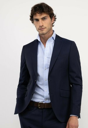 Hombre vestido con un traje azul marino y una camisa de rayas azul claro, de pie frente a un fondo blanco, con las manos en los bolsillos.