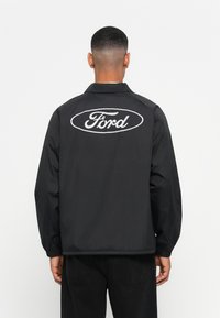 Black windbreaker jacket with a logo on the back. Features a collar, elastic cuffs, and a loose fit. Smooth fabric, minimalistic design.
