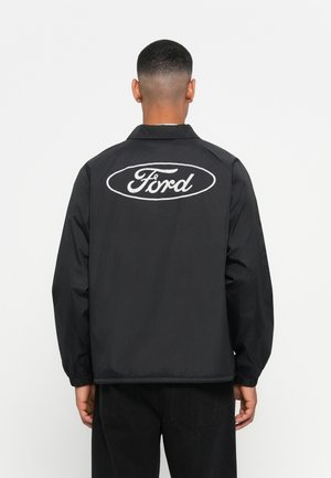 Black windbreaker jacket with a logo on the back. Features a collar, elastic cuffs, and a loose fit. Smooth fabric, minimalistic design.