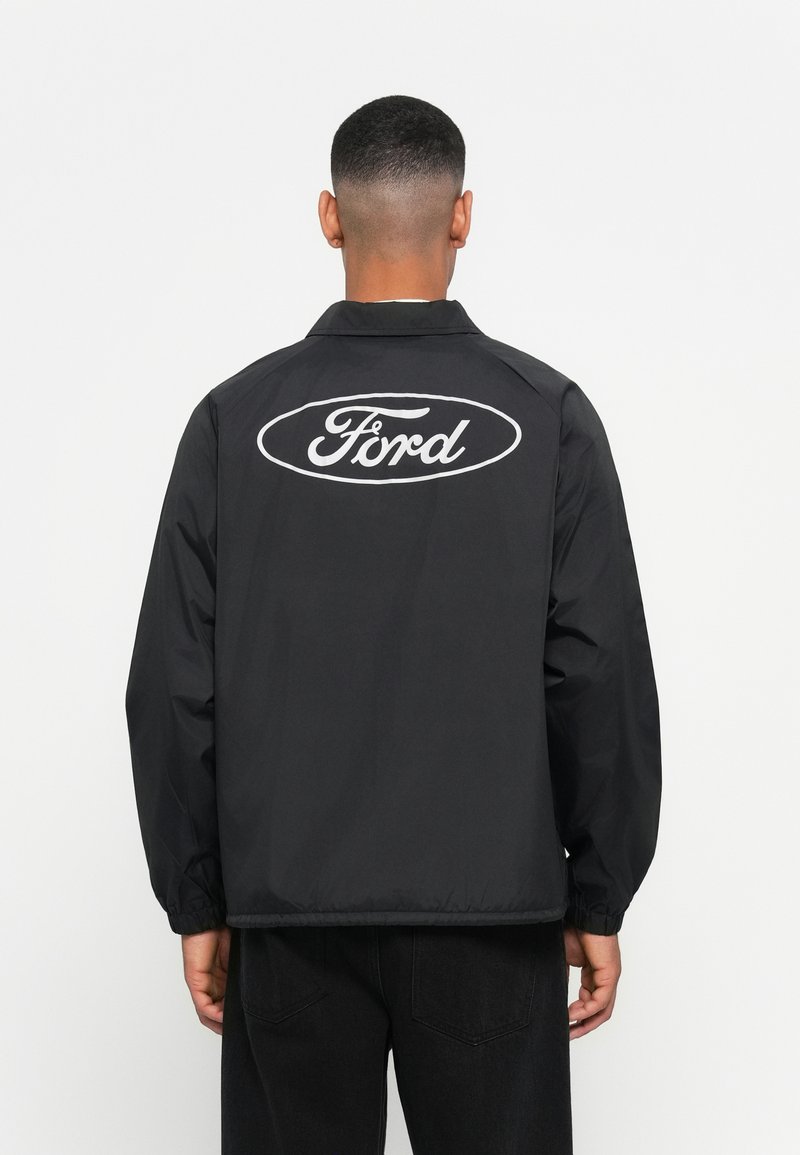 Black windbreaker jacket with a logo on the back. Features a collar, elastic cuffs, and a loose fit. Smooth fabric, minimalistic design.