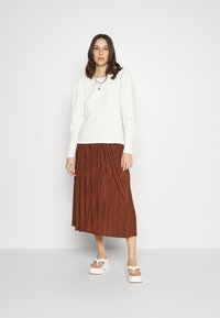 Sweater branco texturizado com mangas longas, combinado com uma saia midi plissada castanha. Modelo usa sandálias de plataforma brancas e colares sobrepostos.