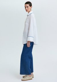 Camisa blanca con ribetes azules, de mangas largas y con un bolsillo frontal, combinada con pantalones de denim azul de pierna ancha y zapatillas de color claro.