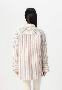 Striped shirt in light tones with red and navy lines, featuring a pointed collar, long sleeves, and a curved hem. Smooth fabric texture.