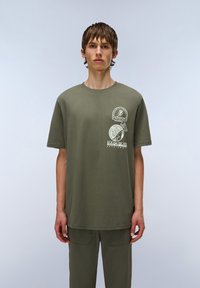 Camiseta de algodón verde oliva con mangas cortas, cuello redondo y gráficos impresos en el pecho que presentan elementos circulares y de texto.