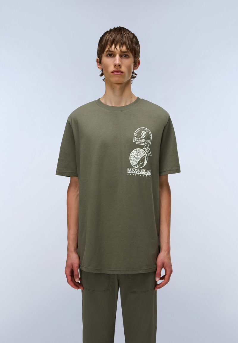 Camiseta de algodón verde oliva con mangas cortas, cuello redondo y gráficos impresos en el pecho que presentan elementos circulares y de texto.