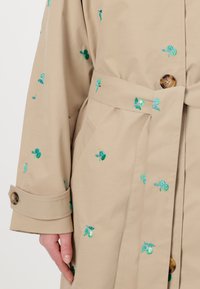 Beige trenchcoat met groene bloemborduursels, voorzien van een strikceintuur, oversized pasvorm en een grote knoopdetail op de manchet.