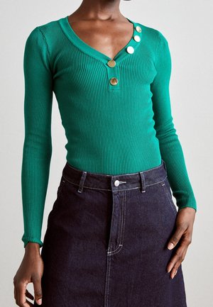 Grøn ribbet langærmet top med V-hals og fire guldknapper kombineret med en høj talje mørk denim nederdel på en model.