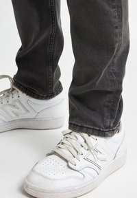 Zapatillas blancas con detalles perforados, cordones en un tono gris claro, combinadas con jeans gris oscuro. El zapato cuenta con una superficie texturizada y un logo en el lateral.