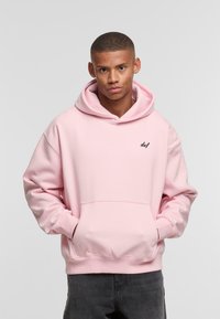 Light pink hoodie made of soft fabric, featuring a kangaroo pocket, ribbed cuffs, and a small black "def" logo on the chest. Grey pants.