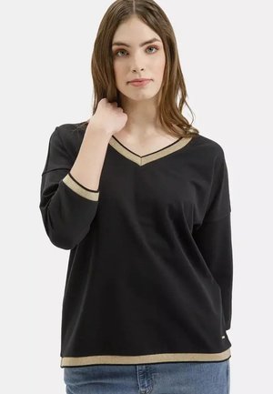 Młoda kobieta ubrana w czarny sweter z dekoltem w serek, wykończony złotą lamówką na kołnierzu, mankietach i u dołu, w połączeniu z niebieskimi dżinsami, na białym tle.