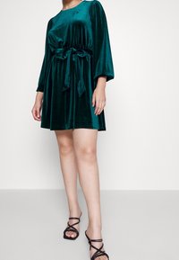 Sammet grön klänning med en figurnära midja och rosett, med puffärmar och en utsvängd kjol. Buren med svarta sandaler med remmar.