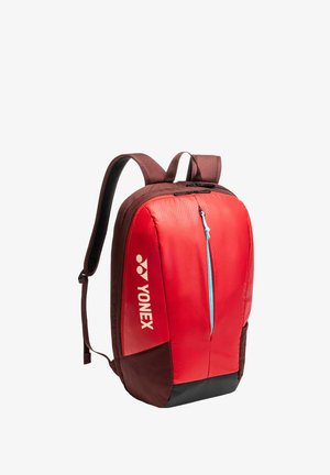 Mochila Yonex en rojo y marrón con tirantes acolchados, bolsillo delantero con cremallera y tirador azul, y gran compartimento principal con cremallera en la parte superior.