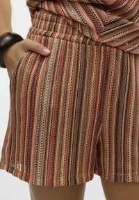 Ręka w kieszeni, nosząc wielokolorowe, prążkowane szorty z brązowymi, pomarańczowymi, czerwonymi i beżowymi odcieniami oraz czarną bransoletkę na nadgarstku.