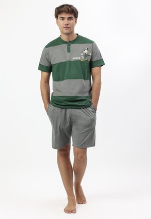 Joven de pie descalzo que lleva una camiseta de manga corta con rayas verdes y grises con un pato de dibujos animados y pantalones cortos grises, con las manos en los bolsillos.