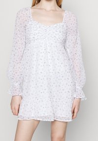 Biała sukienka w kwiaty z długimi, bufkowymi rękawami, z marszczoną górą i warstwowym dołem. Całość pokryta małymi niebieskimi i fioletowymi kwiatkami.