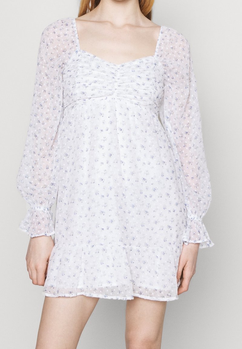 Biała sukienka w kwiaty z długimi, bufkowymi rękawami, z marszczoną górą i warstwowym dołem. Całość pokryta małymi niebieskimi i fioletowymi kwiatkami.