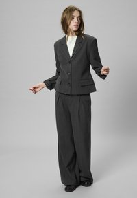 Ženska oblečena v temno sivo oversize suknjo z belo majico in črnimi čevlji, ki pozira proti enobarvni svetlo sivi ozadju.