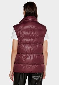 Gilet imbottito bordeaux con colletto alto, design trapuntato e senza maniche. Texture liscia e lucida, indossato sopra una maglietta bianca e pantaloni neri.