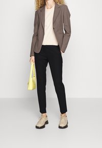 Blazer marron ajusté sur un haut beige clair, pantalon noir et chaussures beiges. Tient un petit sac à main jaune. Design simple et moderne.