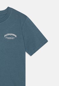 Camiseta de algodón en color azul verdoso con cuello redondo. Presenta mangas cortas y un logo de la marca bordado en blanco en el lado izquierdo del pecho. Textura suave.