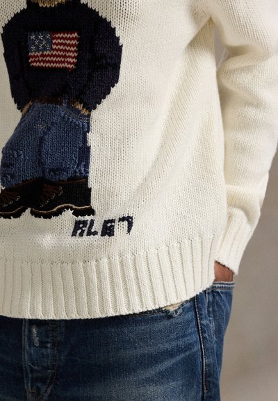 Λευκό πλεκτό πουλόβερ με πολύχρωμο κεντητό σχέδιο μιας φιγούρας με αμερικανική σημαία στο στήθος, συνδυασμένο με μπλε τζιν παντελόνι.