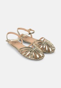 Sandales dorées métalliques à lanières avec une semelle plate, un design à bout ouvert et une bride de cheville. Présente des détails entrecroisés sur le devant.