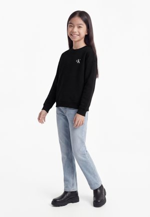 Jeune fille souriante debout portant un sweat-shirt noir avec le logo blanc CK, un jean bleu clair et des bottes noires épaisses sur fond blanc.