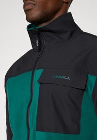 Chaqueta con cremallera en negro y verde azulado, que presenta un material texturizado similar al forro polar y un bolsillo en el pecho con cremallera que incluye un detalle del logotipo. Diseño de cuello alto.