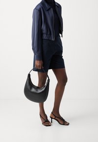 Musta nahkalaukku, jossa on kaareva muoto, teksturoitu pinta ja lyhyet kahvat. Yhdistetty tummansinisellä lyhyellä takilla, tummansinisillä shortseilla ja mustilla korollisandaaleilla.