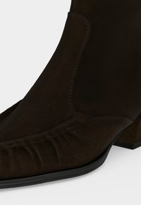 Bottine en daim marron foncé à bout carré, talon bloc bas et détails cousus sur les panneaux avant et latéraux.