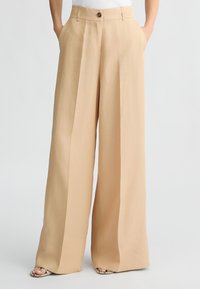 Pantaloni beige a gamba larga realizzati in tessuto morbido, con bottone frontale, tasche laterali e vestibilità ampia che si allarga all'orlo.