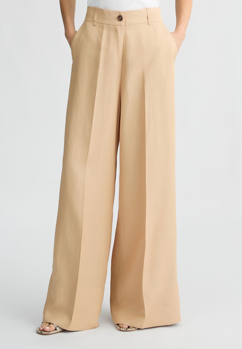 Pantaloni beige a gamba larga realizzati in tessuto morbido, con bottone frontale, tasche laterali e vestibilità ampia che si allarga all'orlo.
