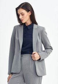 Szary marynarka o dopasowanym kroju z wyraźnymi ramionami, klapami i gładką teksturą. Połączona z ciemnoniebieską koszulą z kołnierzykiem. Widoczne subtelne prążki.