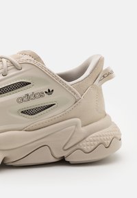 Beige sneaker med ovandel i mocka och mesh, uttalad sula och Adidas-logga på sidan. Har texturerade detaljer och en unik siluett.