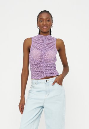 Mladá žena s pletenými vlasmi, ktorá nosí kruhové náušnice, fialový vrchný diel bez rukávov so záhybmi a svetlomodré rifle s vysokým pásom, usmieva sa s rukou v vrecku.