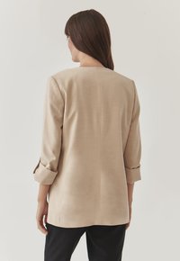 Beige sakový blejzr s rovnými liniemi, rukávy po lokty vyhrnuté, a hladkým povrchem. Nositelný přes černé kalhoty.