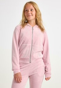 Nuori nainen, joka pukee ylleen vaaleanpunaisen velourista valmistetun vetoketjuhupullisen takin ja siihen sopivat housut, seisoo tasaisella valkoisella taustalla.
